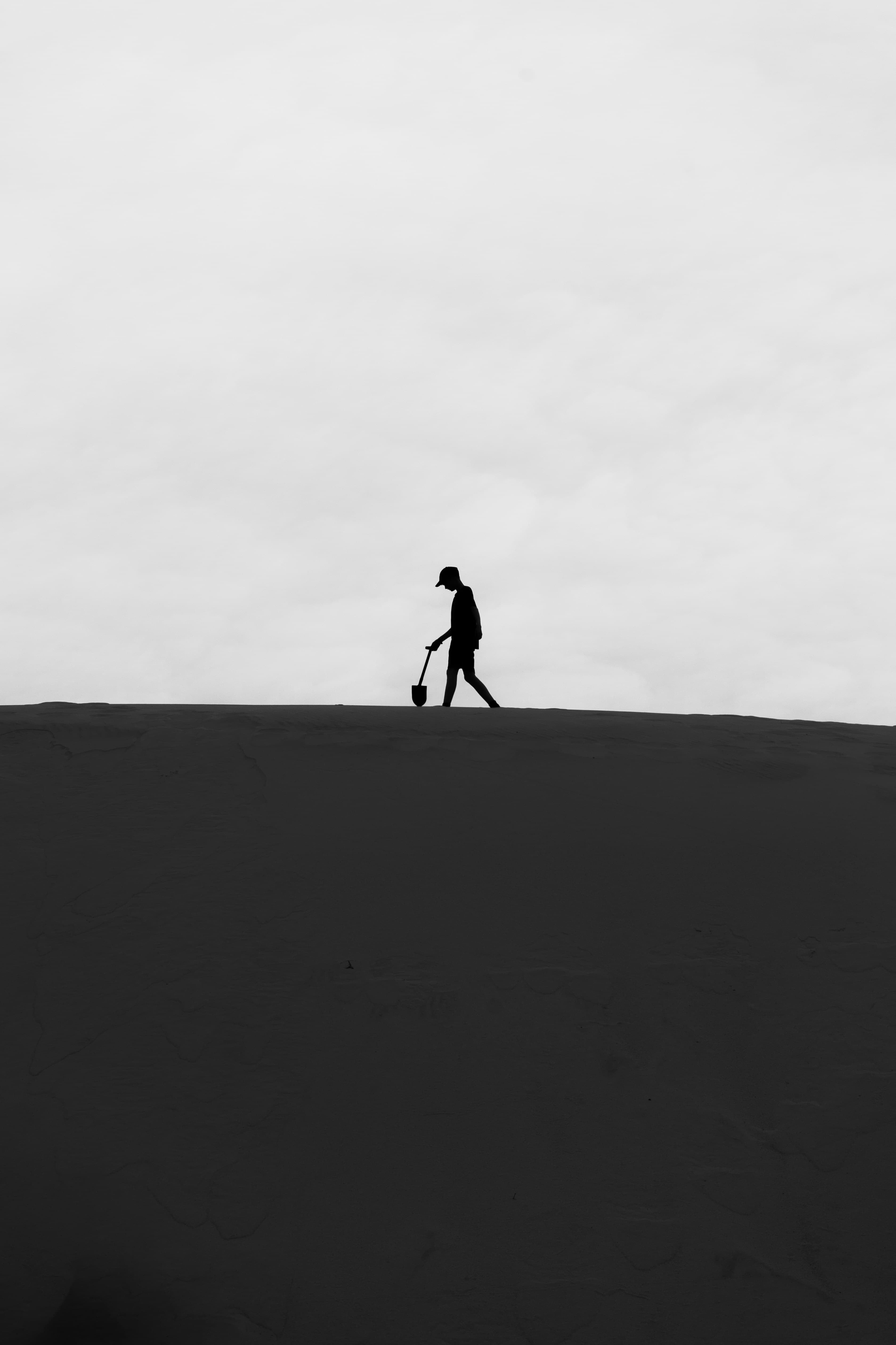 Silhouette of a person walking on sand dunes with a metal detector under a cloudy sky.